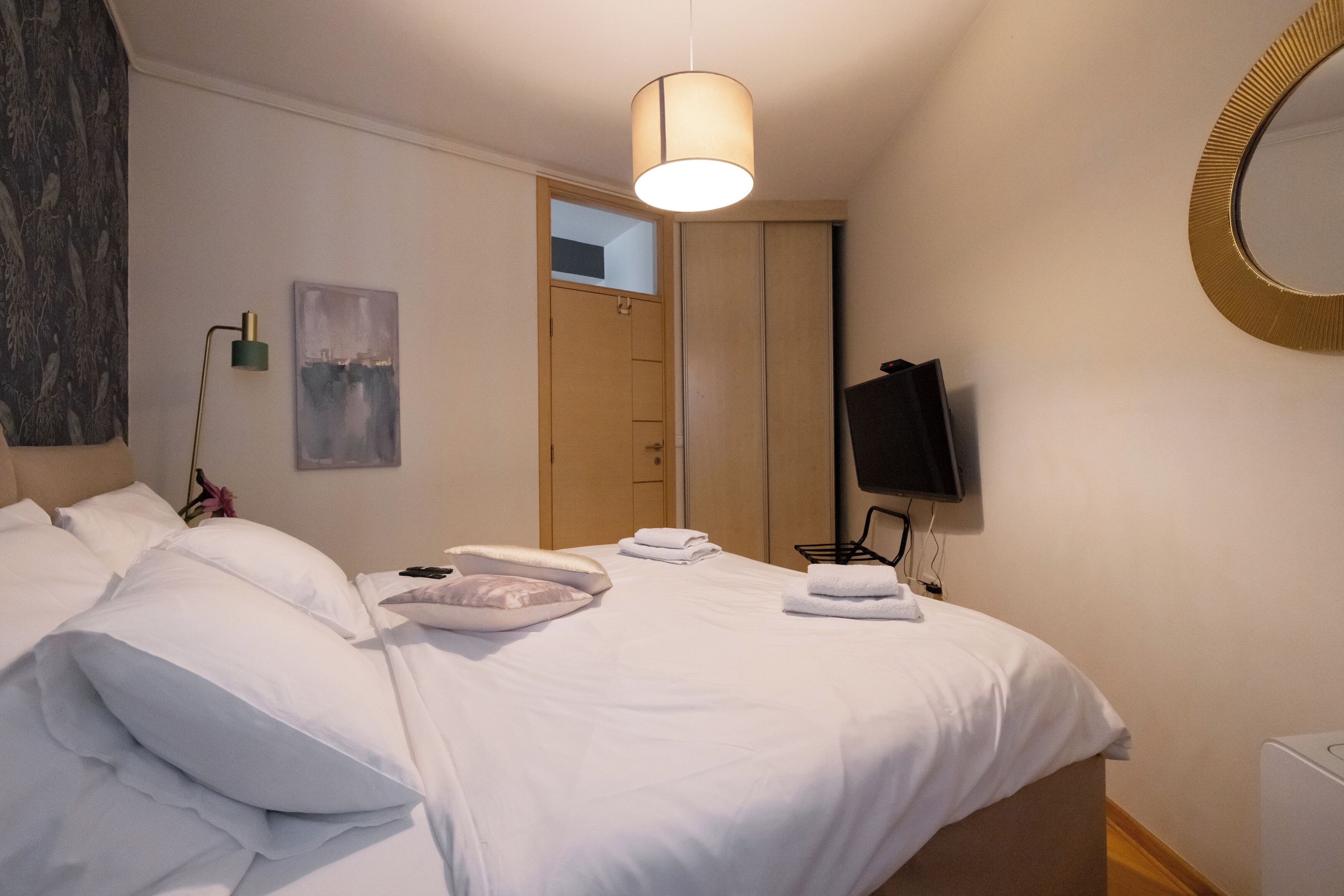 Een netjes opgemaakt bed met witte lakens en kussens, een bedlamp, een flatscreen-televisie en een ingelijste foto aan de muur.