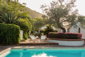 Un espace piscine avec des chaises longues, un bâtiment blanc et une végétation luxuriante.