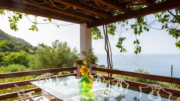 Une table à plateau en verre, avec des chaises et un vase de fleurs, placée devant un décor pittoresque composé de verdure et de mer.
