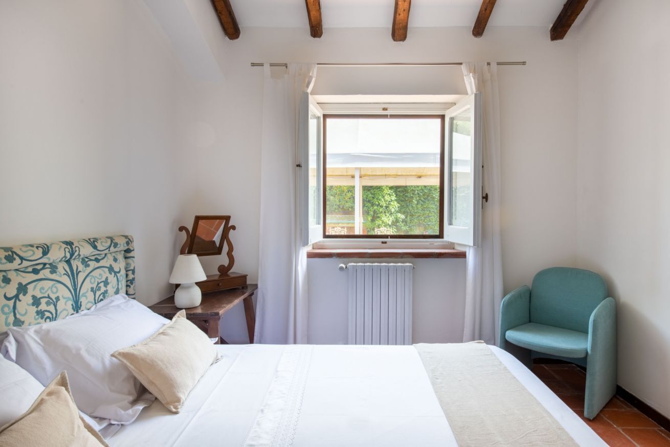 Un dormitorio con cama, ventana, silla y mesita de noche con lámpara.