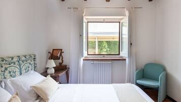 Un dormitorio con cama, ventana, silla y mesita de noche con lámpara.