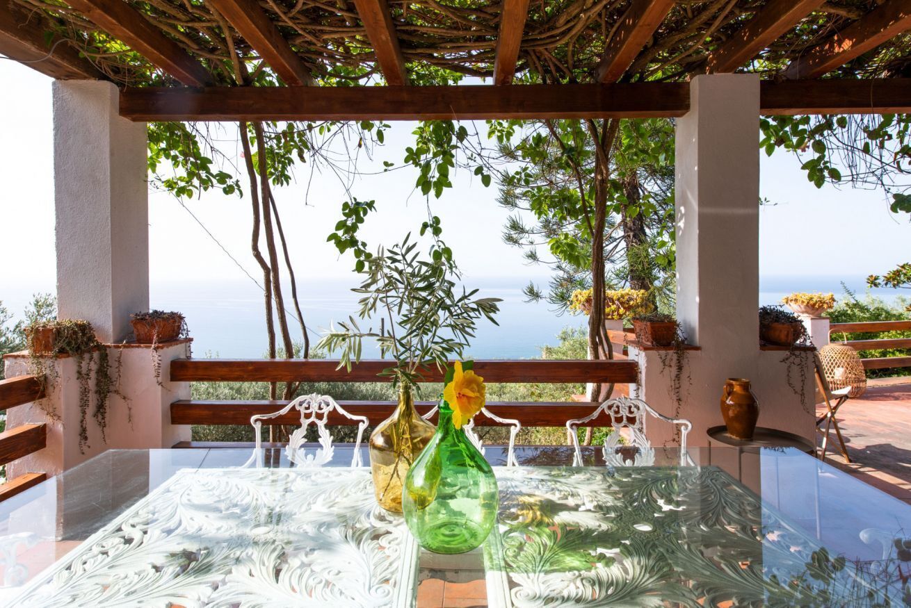 Een tafel met een glazen blad, omringd door witte metalen stoelen, onder een pergola met uitzicht op zee.