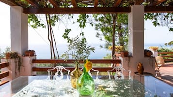 Een tafel met een glazen blad, omringd door witte metalen stoelen, onder een pergola met uitzicht op zee.