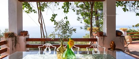 Une table dont le plateau est en verre, entourée de chaises en métal blanc, placée sous une pergola d’où l’on a vue sur la mer.