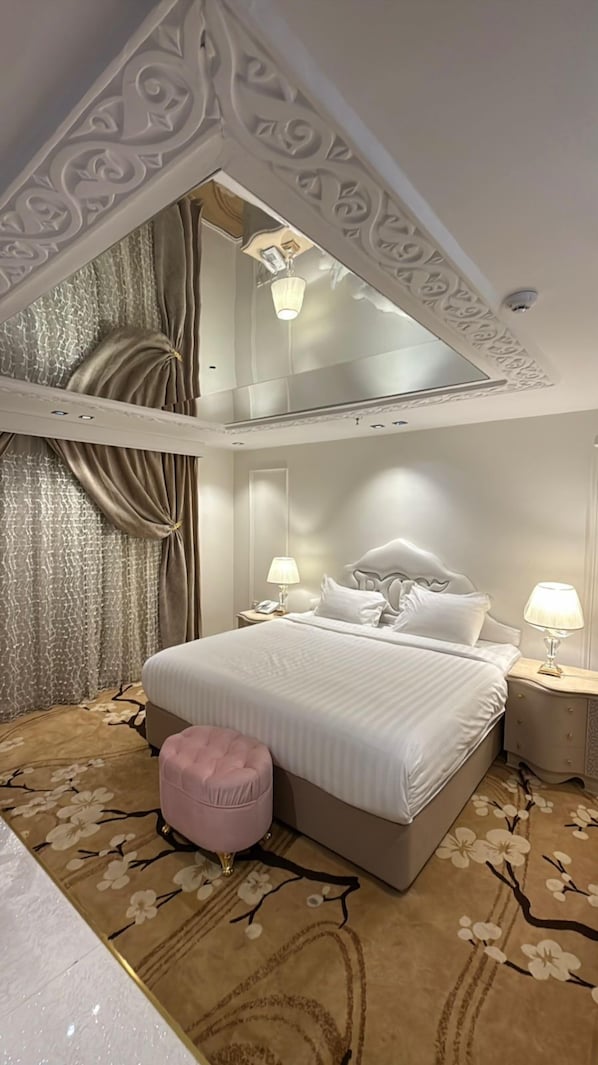 A bedroom with a large bed, two bedside tables, a pink ottoman, and a chandelier.