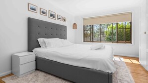 A modern bedroom with a grey upholstered bed, white bedding, a small bedside table, and a large window with a view of greenery. - Verzelha by the Sea - Spacious 3BR Family Stay in Culburra (Culburra Beach)