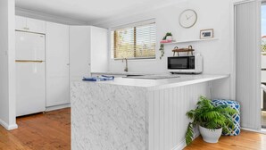 A modern kitchen with white cabinetry, a marble countertop, a built-in microwave, and a window with blinds. - Verzelha by the Sea - Spacious 3BR Family Stay in Culburra (Culburra Beach)