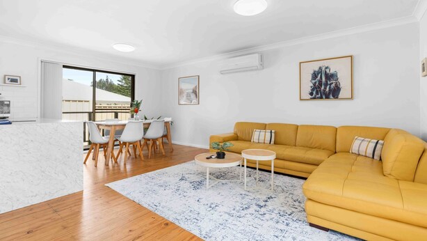 A modern living room with a yellow sofa, a dining area with white chairs, and a large window providing a view of the outdoors. - Verzelha by the Sea - Spacious 3BR Family Stay in Culburra (Culburra Beach)