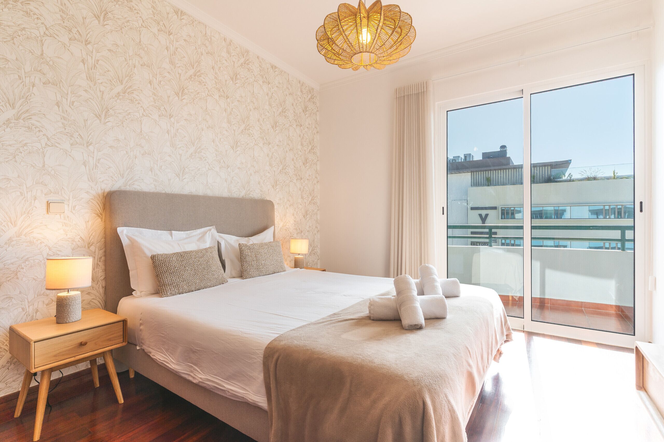 Une chambre d’hôtel avec un grand lit, une table de chevet avec une lampe, une vue sur la ville par une porte coulissante et un balcon avec une balustrade.