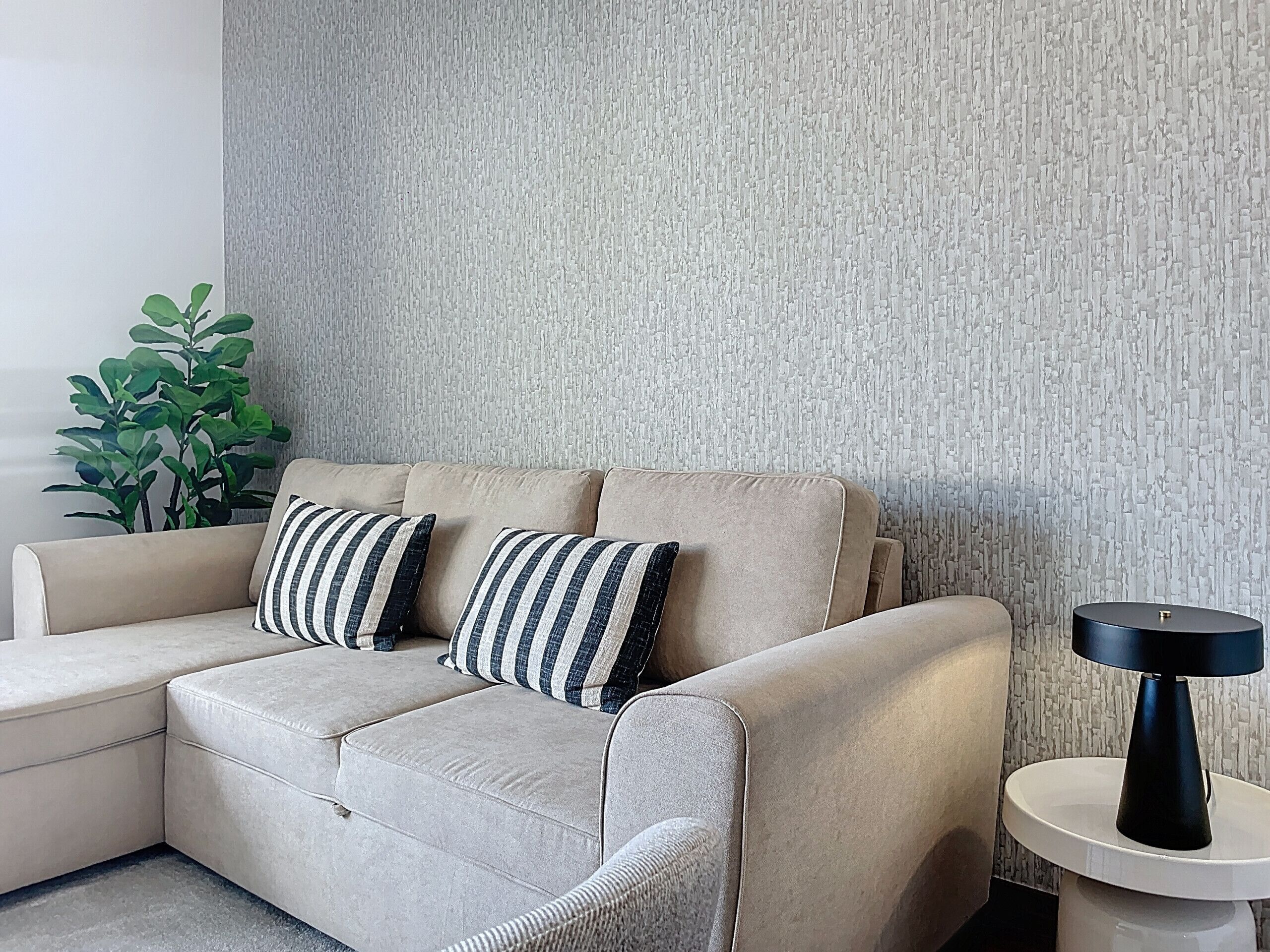 Un canapé d’angle beige agrémenté de coussins à rayures, adossé à un mur texturé, une petite table ronde avec une lampe noire et une plante verte sur le côté gauche.