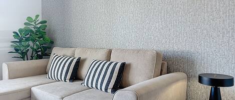 Un canapé d’angle beige agrémenté de coussins à rayures, adossé à un mur texturé, une petite table ronde avec une lampe noire et une plante verte sur le côté gauche.