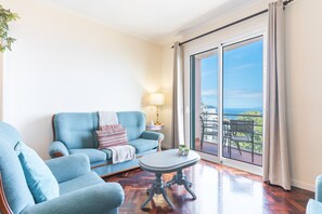 A living room with a sofa, armchair, and round table, a lamp, and a view through a sliding glass door to the outdoors. - Forum Delights Apartment by Atlantic Holiday (Funchal)