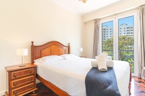 Ein Hotelzimmer mit einem hölzernen Bett, einem Nachttisch mit Lampe und Blick auf Gebäude durch das Fenster.