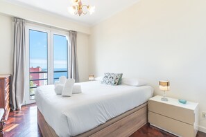 A bedroom with a large bed, a nightstand with a lamp, a dresser, and a view of the sea through the window. - Forum Delights Apartment by Atlantic Holiday (Funchal)