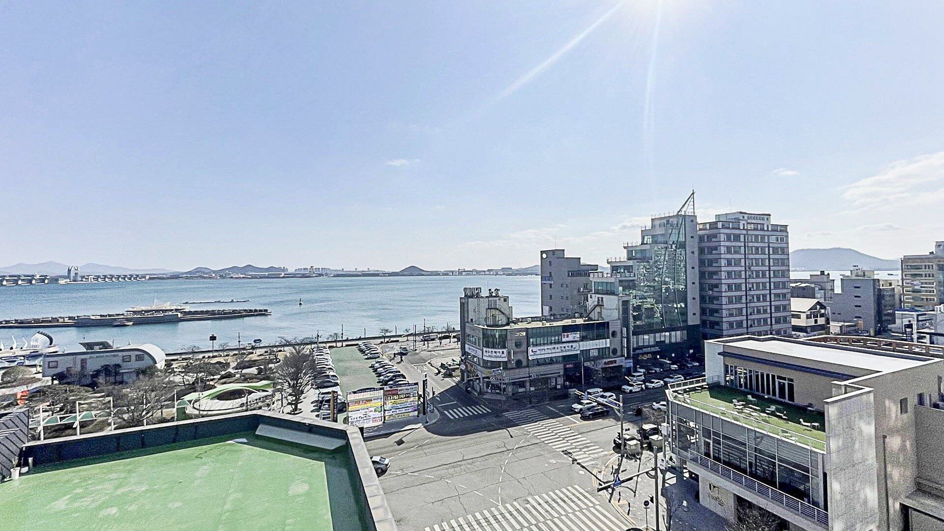 A cityscape with a view of the sea, featuring modern buildings, a parking lot, and a green-roofed structure.