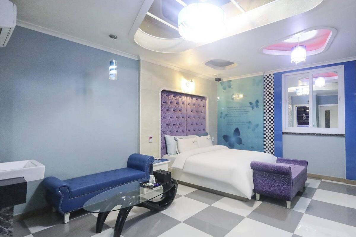 Ein modernes Schlafzimmer mit Schachbrettmuster-Boden, einem Bett mit gestepptem Kopfteil, einem blauen Sofa, einem violetten Hocker und einem Glastisch.