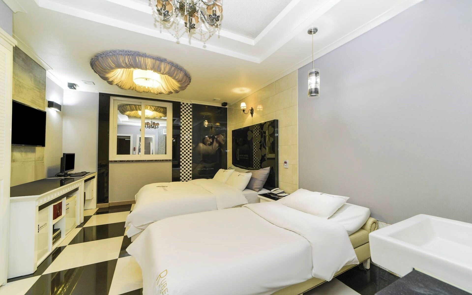 A hotel room with two beds, a desk with a computer, a large mirror, and a chandelier.