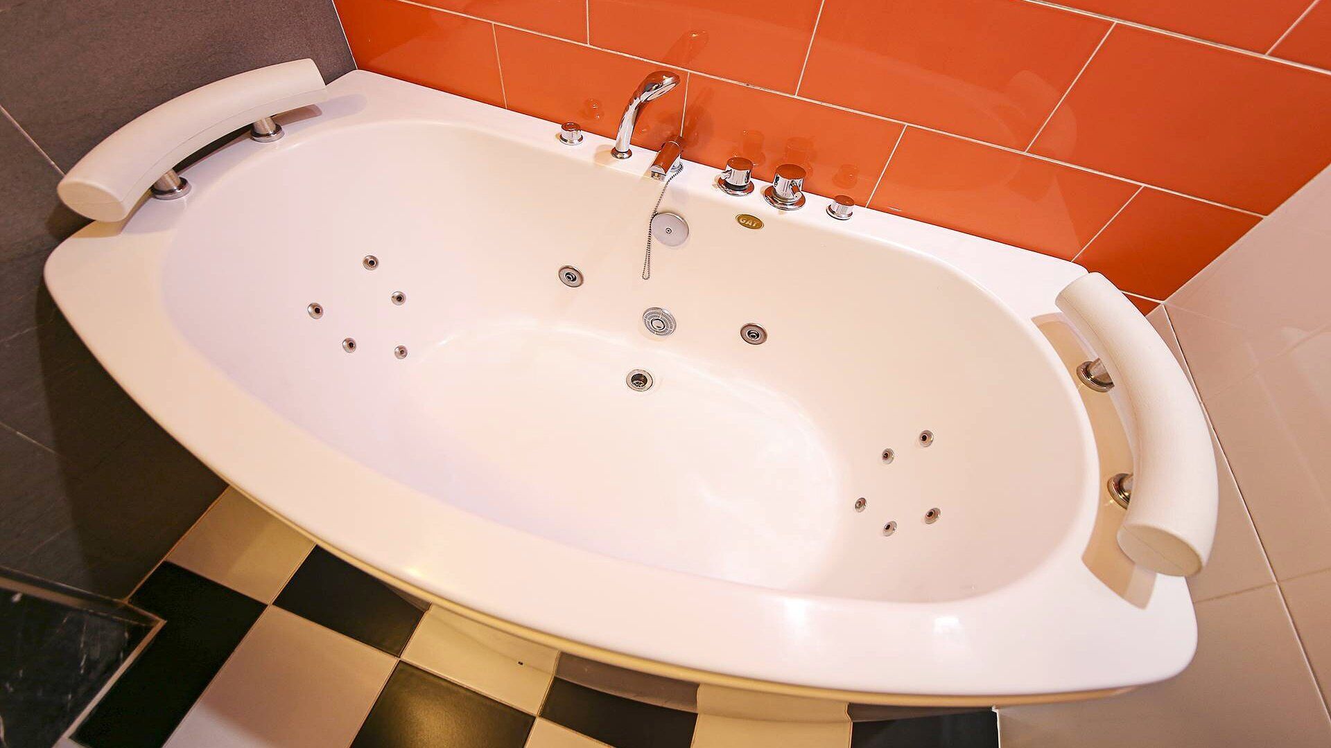 A white freestanding bathtub with multiple faucets and a handheld showerhead, set against a checkered floor and orange-tiled walls.