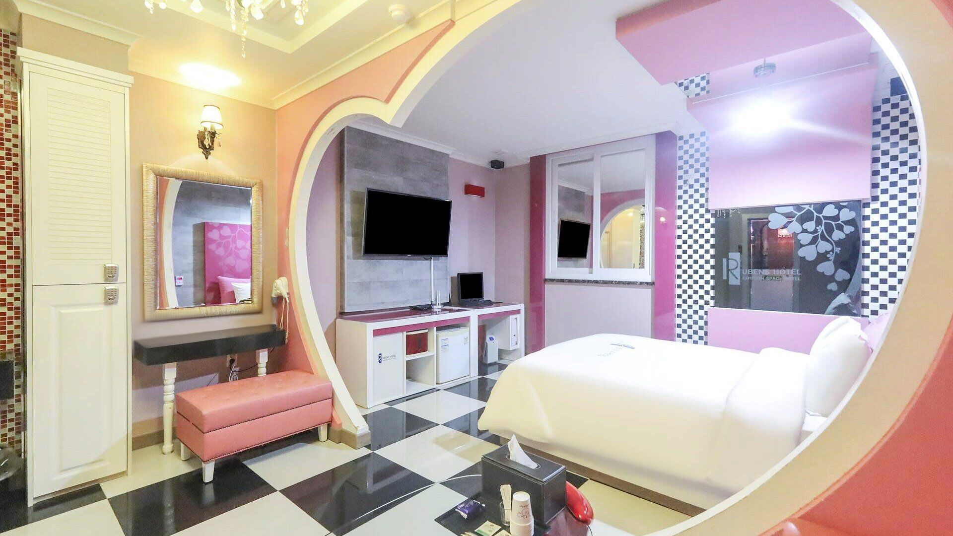 A modern hotel room with a checkered floor, a bed, a vanity with a mirror, a television, and a minibar.