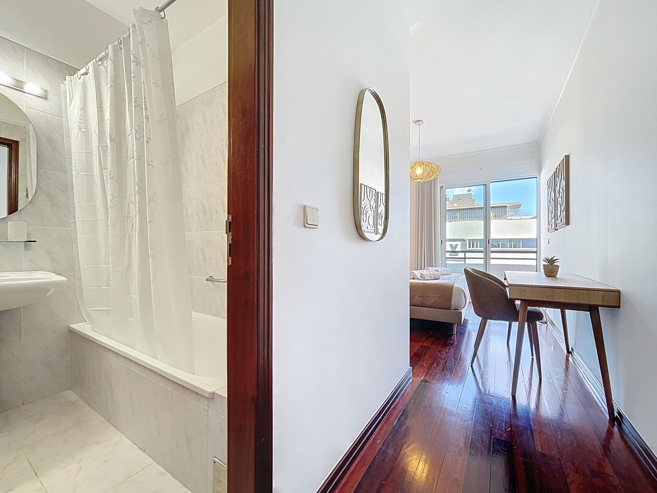 Un baño moderno con ducha, lavamanos y espejo. Se ve una puerta de madera que conduce a un dormitorio con cama, una silla y una mesa. La habitación tiene un ventanal y vistas al exterior.