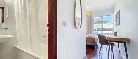Un baño moderno con ducha, lavamanos y espejo. Se ve una puerta de madera que conduce a un dormitorio con cama, una silla y una mesa. La habitación tiene un ventanal y vistas al exterior.