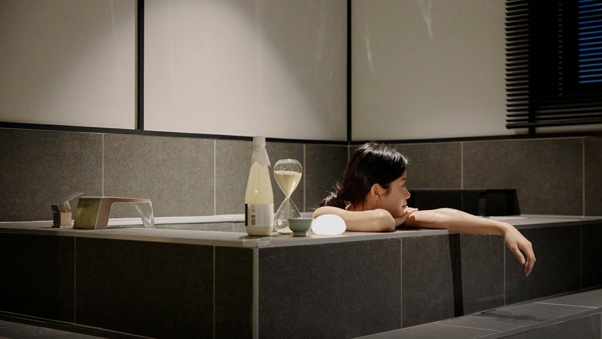 A person resting in a modern bathroom with a bathtub, a bottle, and an hourglass on the counter.