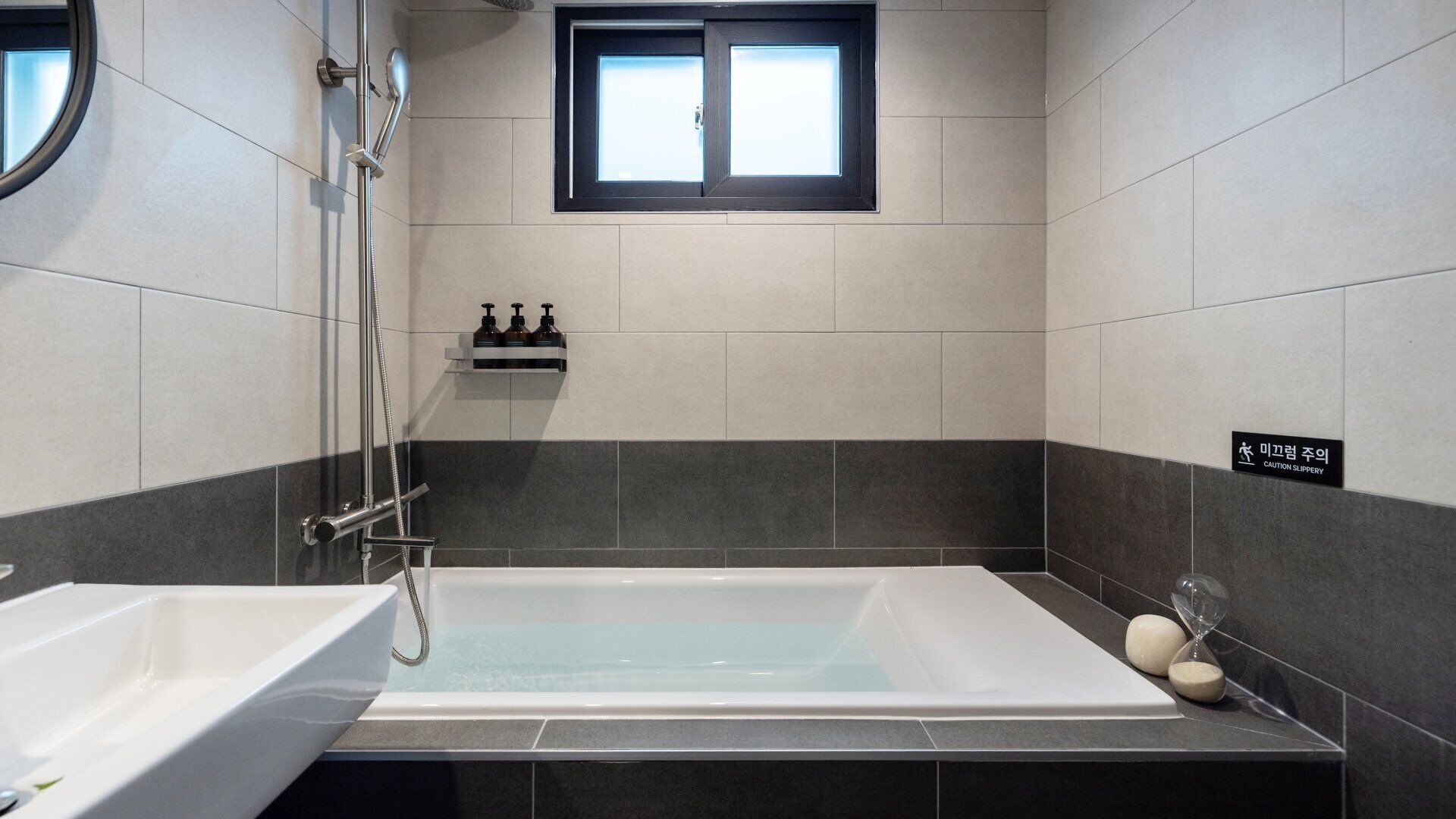 Un baño moderno con bañera, ducha y lavamanos. Hay una ventana sobre la bañera y un letrero de precaución en la pared.