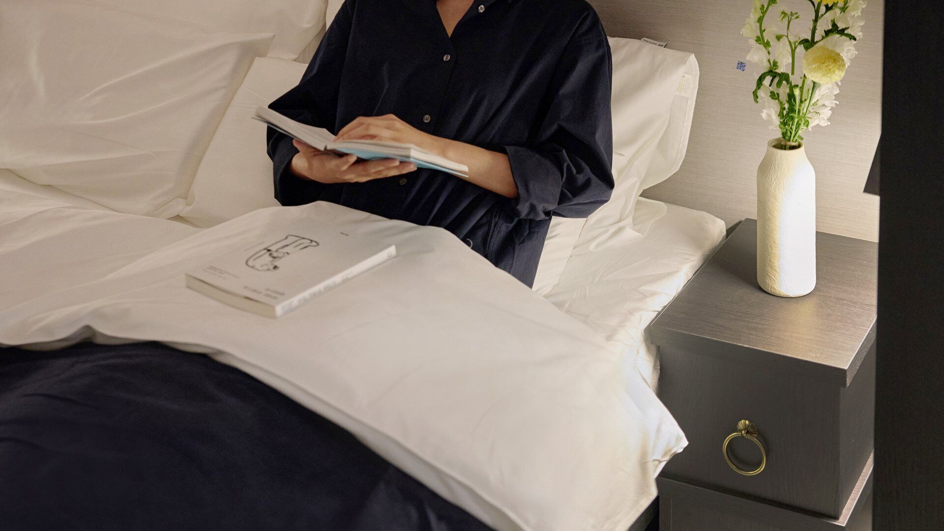 A person reading a book on a bed with white bedding, a white vase with flowers on a bedside table, and a gray nightstand.