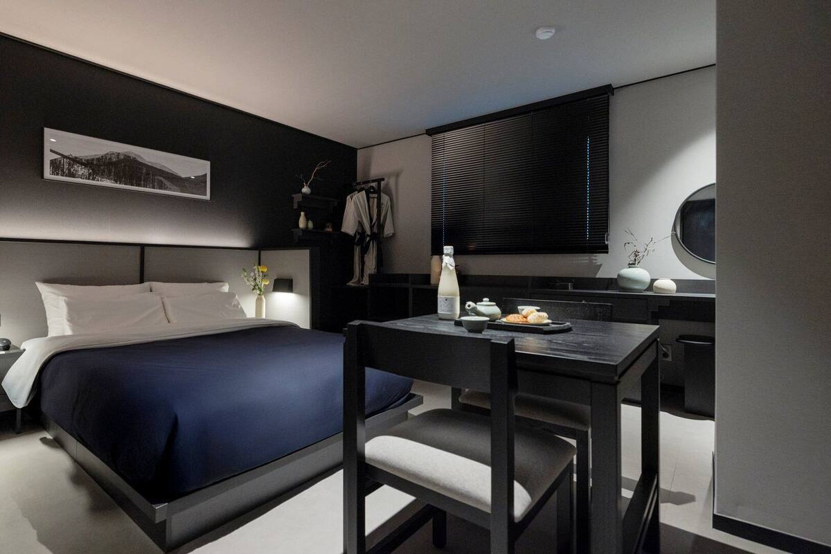 Ein modernes Hotelzimmer mit einem großen Bett, einem Schreibtisch mit Stuhl, einem Nachttisch, einem Fenster mit Jalousien und einem an der Wand hängenden Kunstwerk.
