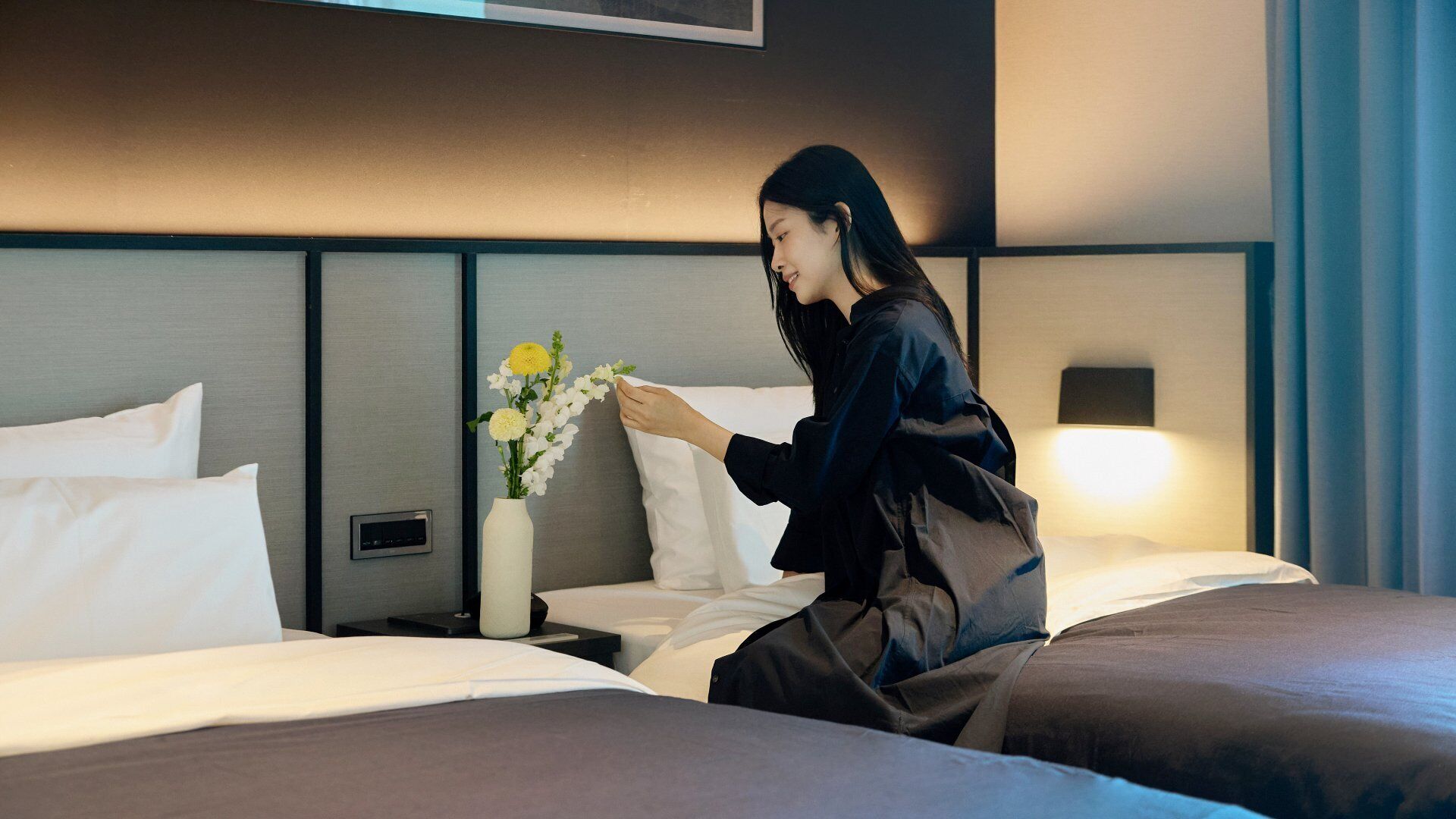 A hotel room with a bed, a bedside table with a vase of flowers, and a control panel on the wall.