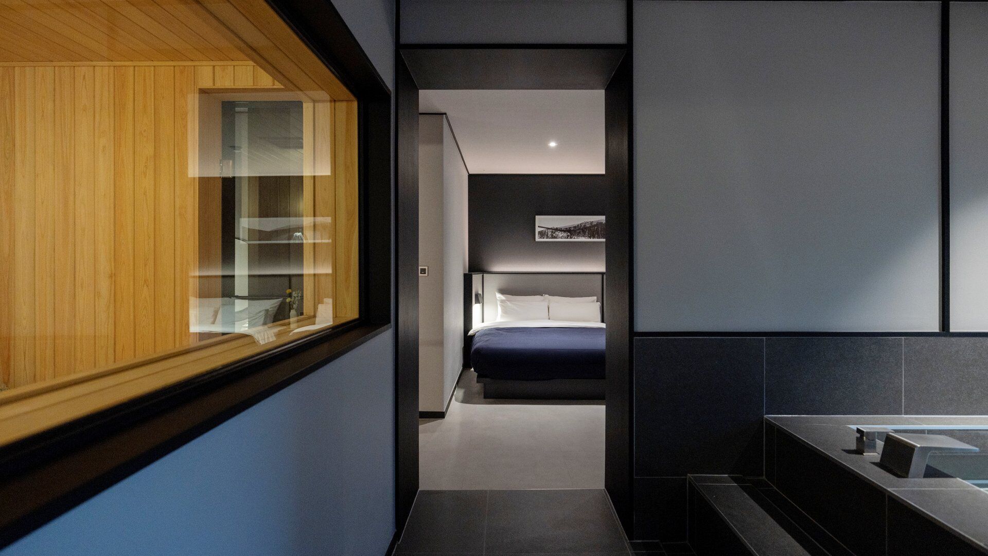 Ein modernes Hotelzimmer mit einem raumhohen Holzfenster, einem Bett mit dunkelblauer Bettdecke, einem Nachttisch und einer Wandlampe.
