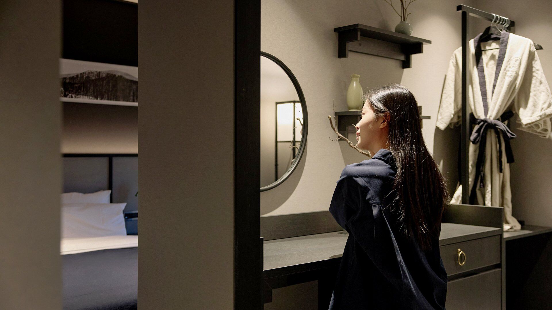 A hotel room with a bed, a mirror, a dresser, and a robe hanging on a rack.