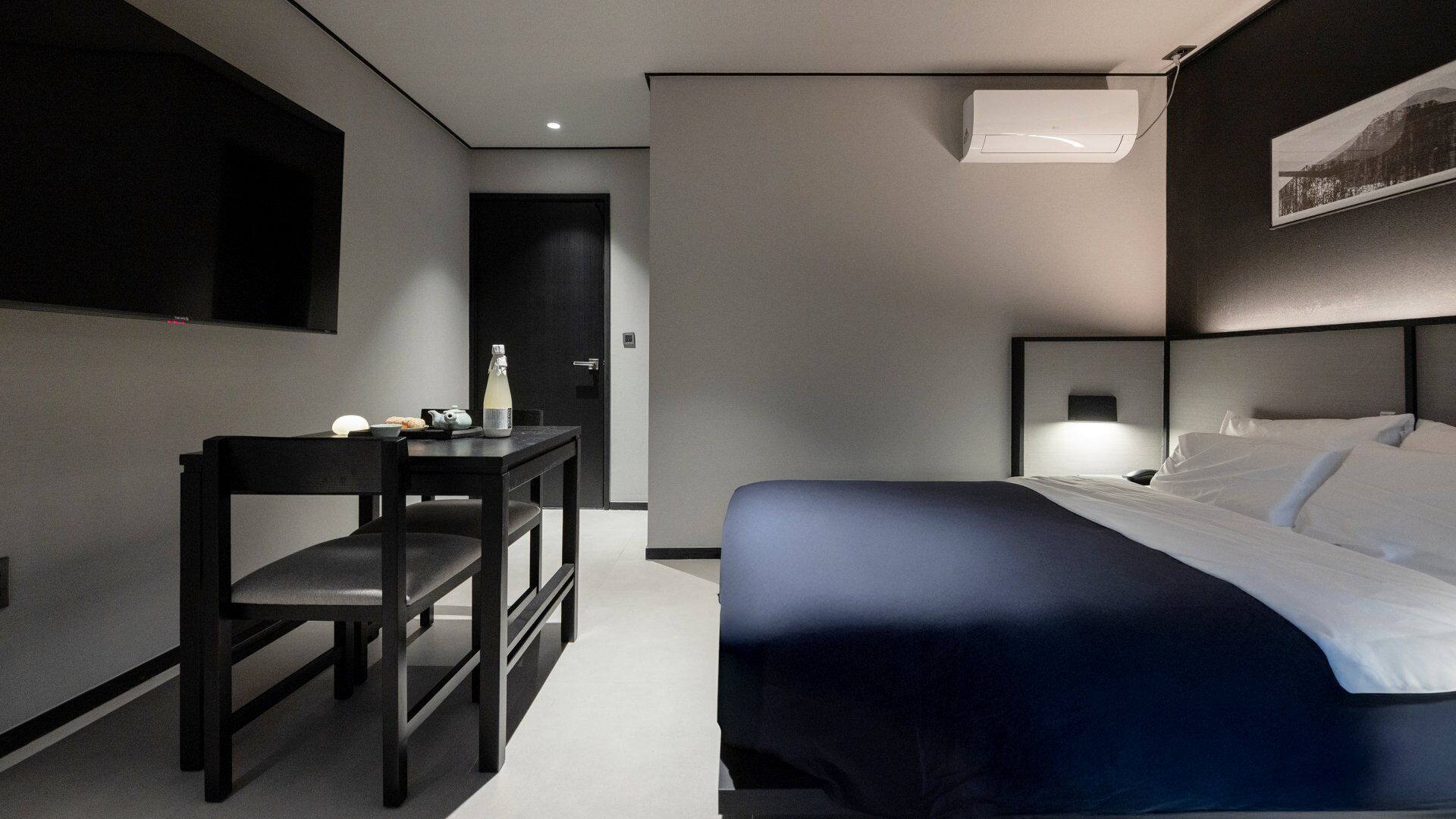 Habitación de hotel moderna con cama, escritorio y silla. Tiene aire acondicionado de pared y un cuadro enmarcado sobre la cama.