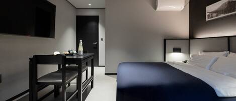 A modern hotel room with a bed, a desk, and a chair. There is a wall-mounted air conditioner and a framed picture above the bed.