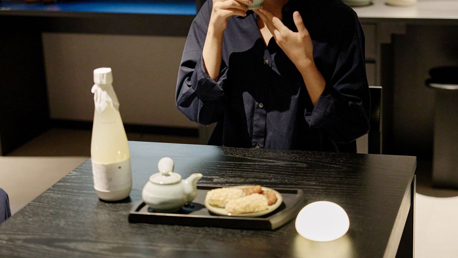 A person standing at a table with a bottle, a teapot, and a plate of food.