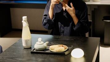 A person standing at a table with a bottle, a teapot, and a plate of food.