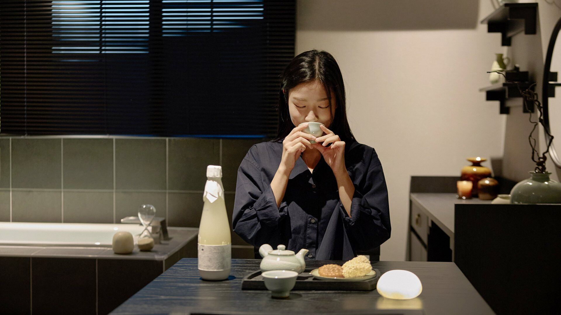 A person seated at a table with a teapot, a bottle, and a plate of food.