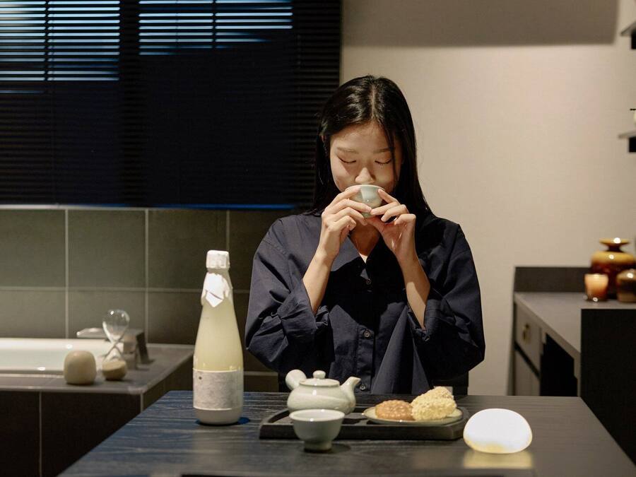 A person seated at a table with a teapot, a bottle, and a plate of food.