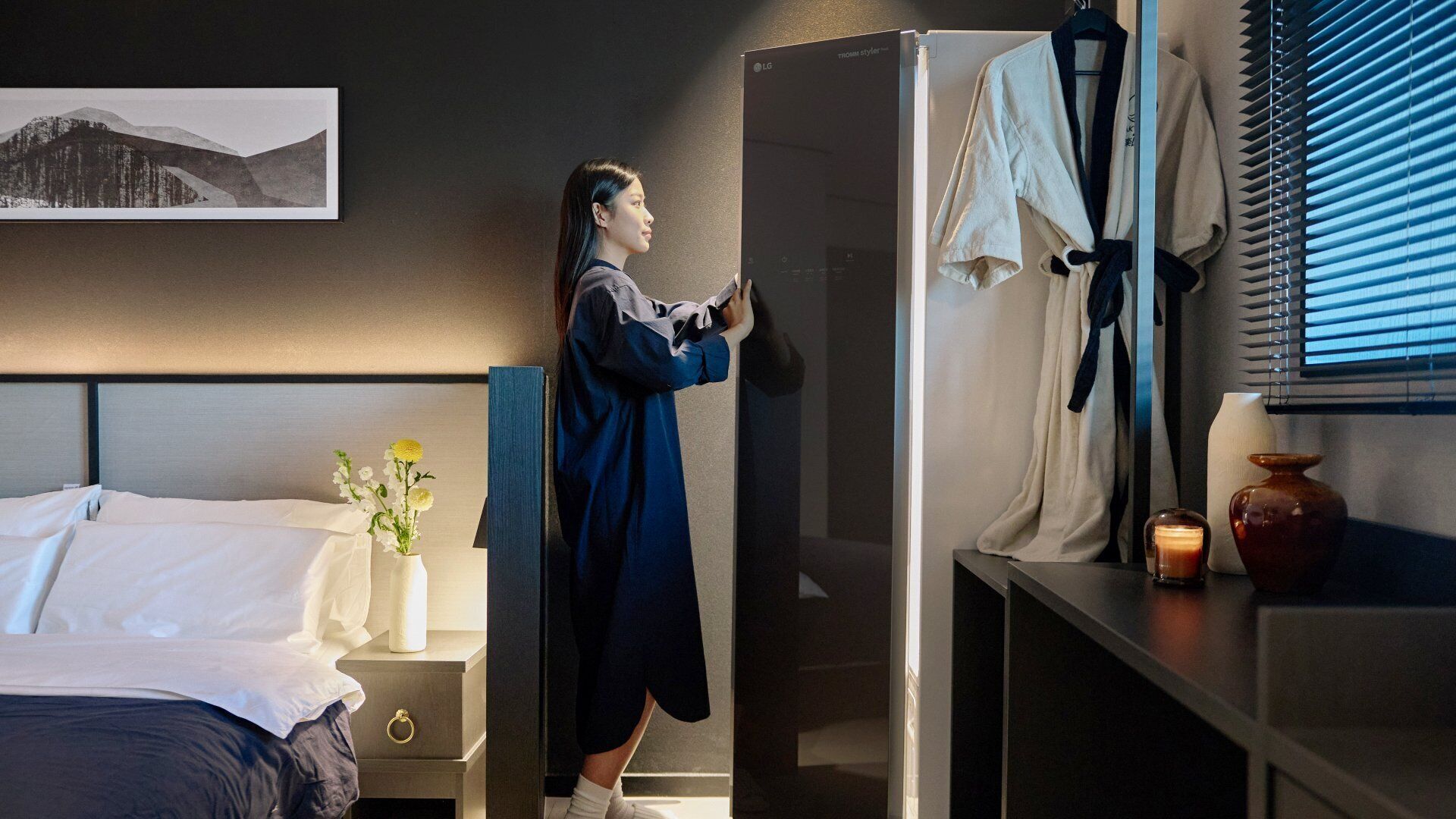 A hotel room with a bed, a nightstand with a vase of flowers, a dresser, and a window with blinds.
