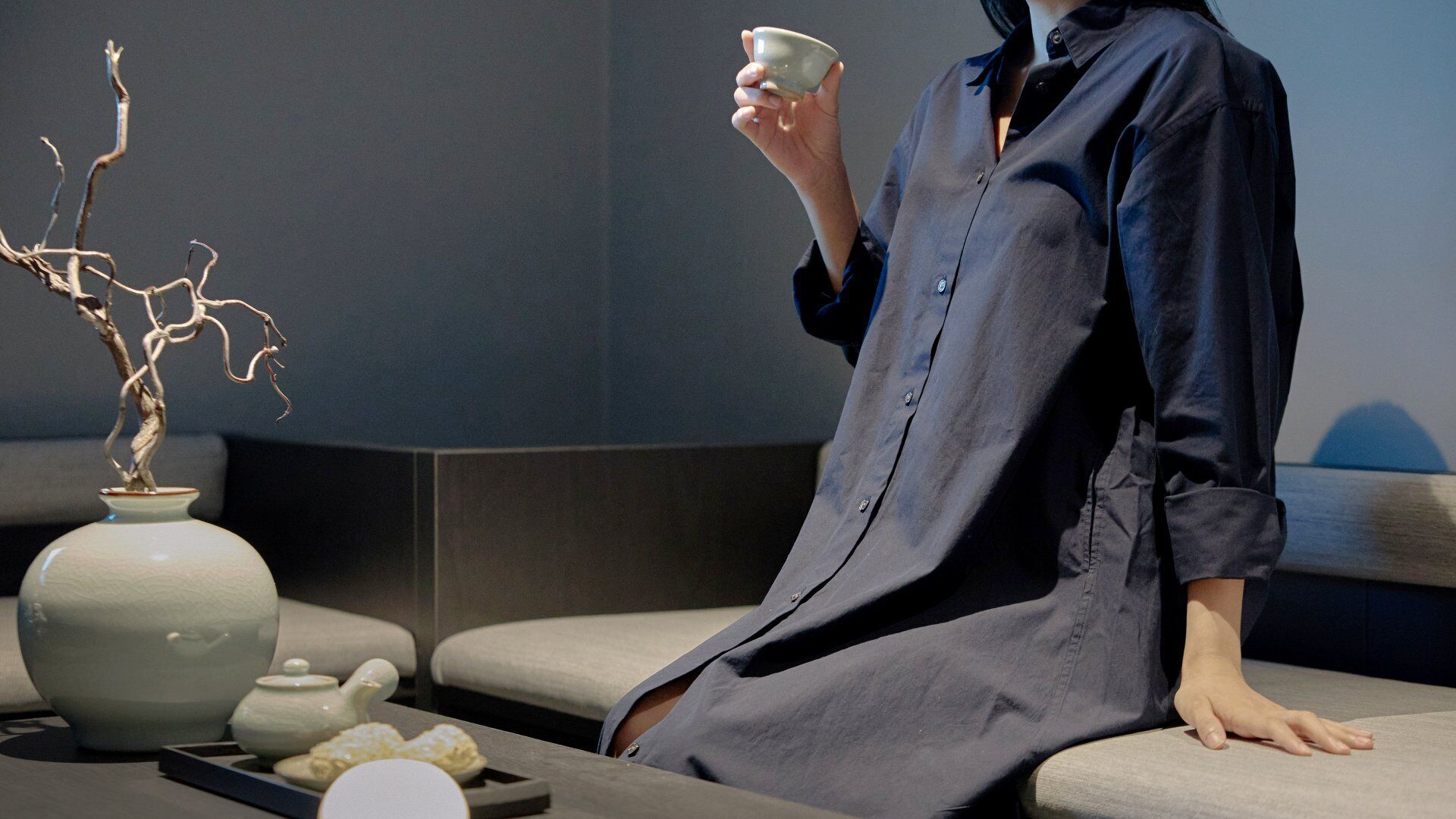 Une personne assise sur un canapé dans un intérieur moderne, avec un vase et une théière posés sur une table.