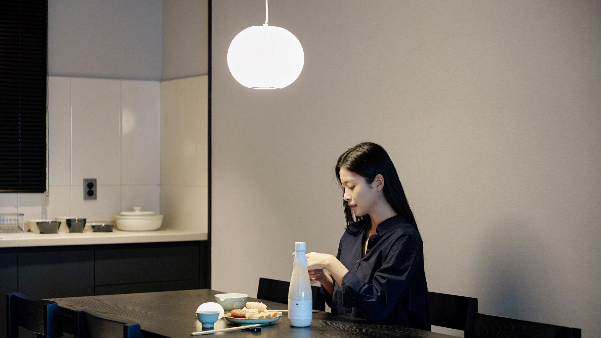 Une personne est assise à une table, devant une bouteille, des bols et une assiette de nourriture, dans une cuisine moderne.