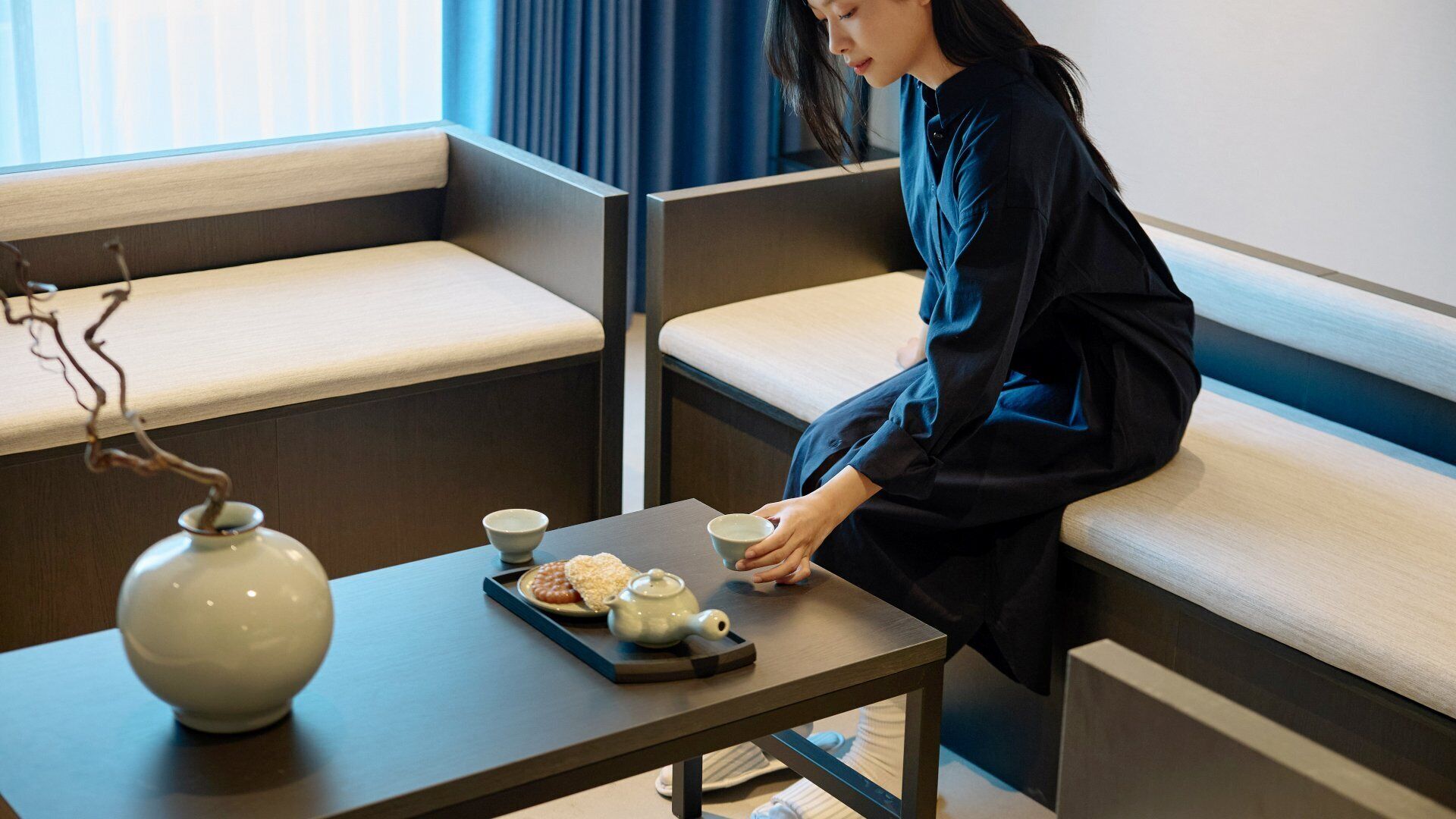 A person seated in a modern hotel room with a sofa and a small table with a teapot and a bowl.