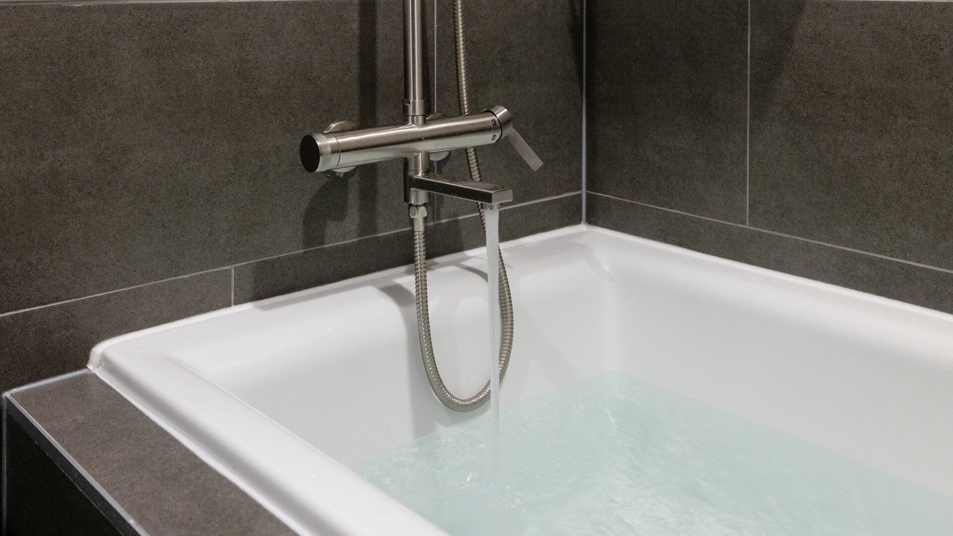 A modern bathroom with a white bathtub, chrome fixtures, and grey tiled walls.