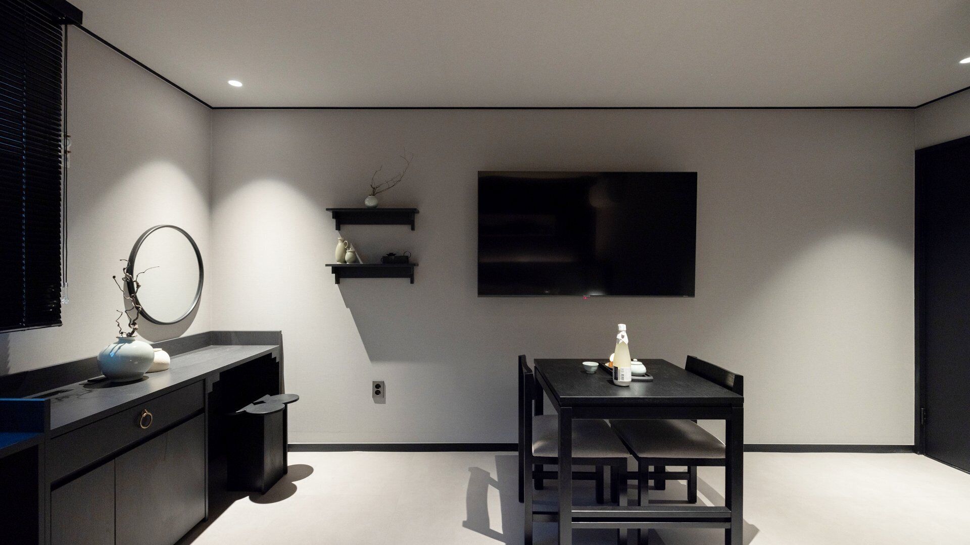 A modern, minimalist room with a dark wooden table and chairs, a large flat-screen TV mounted on the wall, and a small shelf with decorative items.