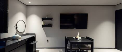 A modern, minimalist room with a dark wooden table and chairs, a large flat-screen TV mounted on the wall, and a small shelf with decorative items.