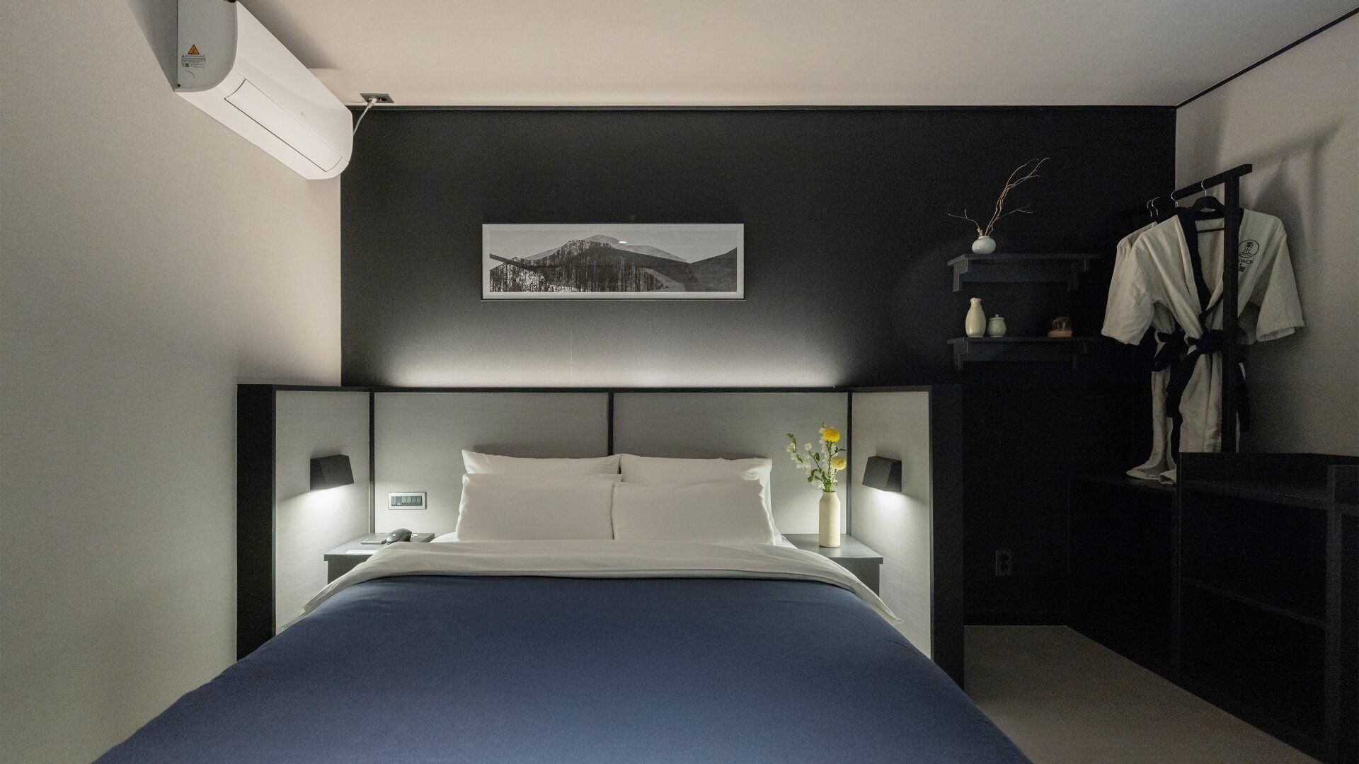 A modern bedroom with a large bed, bedside lamps, a framed picture on the wall, and a dark nightstand with a vase of flowers.