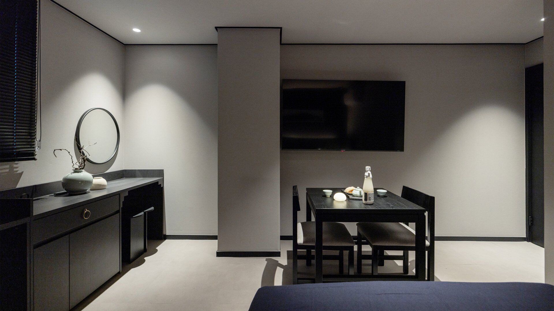 A modern bedroom with a bed, a bedside table with a mirror, a dresser, a TV mounted on the wall, and a dining table with chairs.