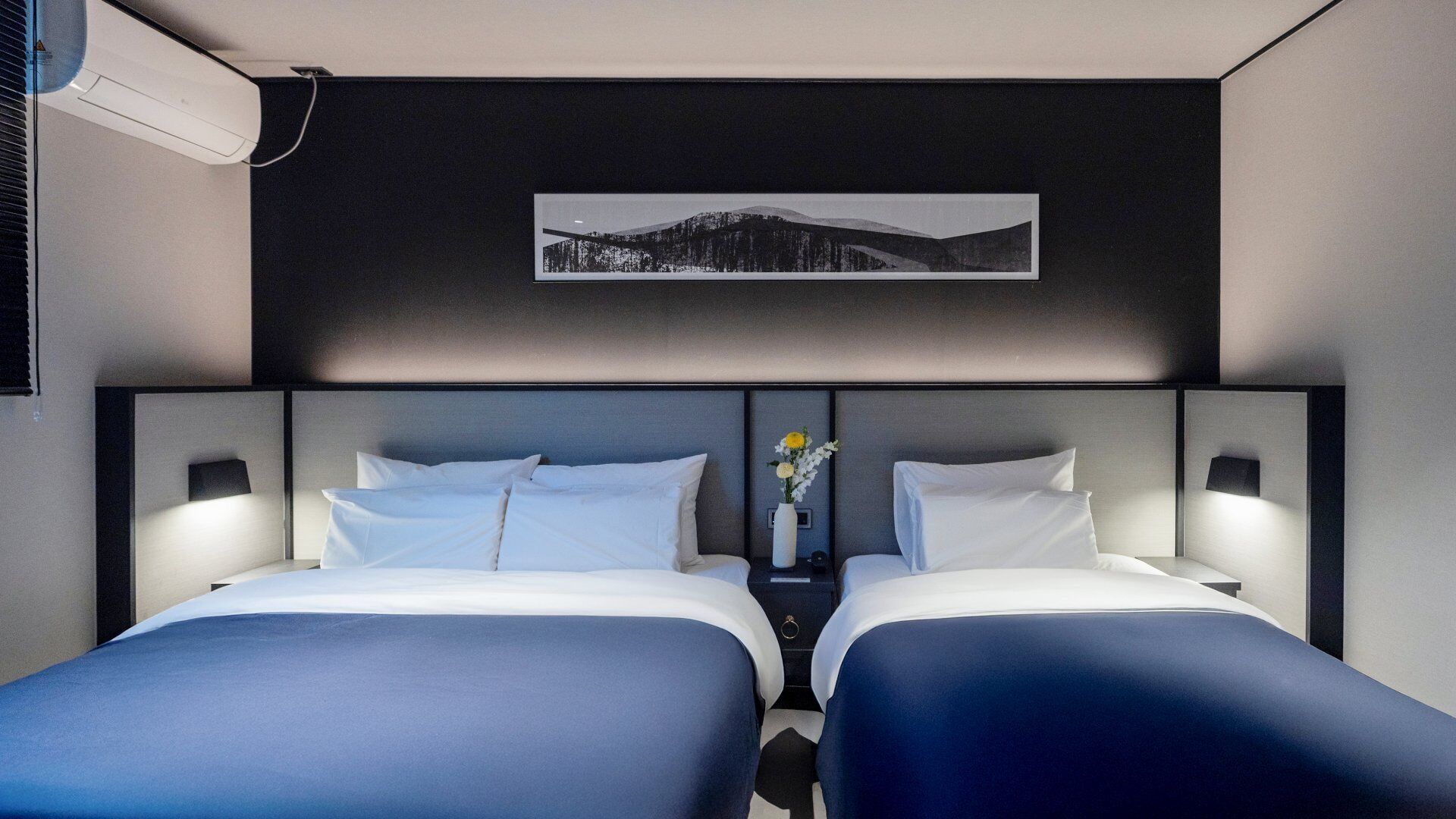 A hotel room with two beds, a nightstand with a vase of flowers, and a framed picture on the wall.