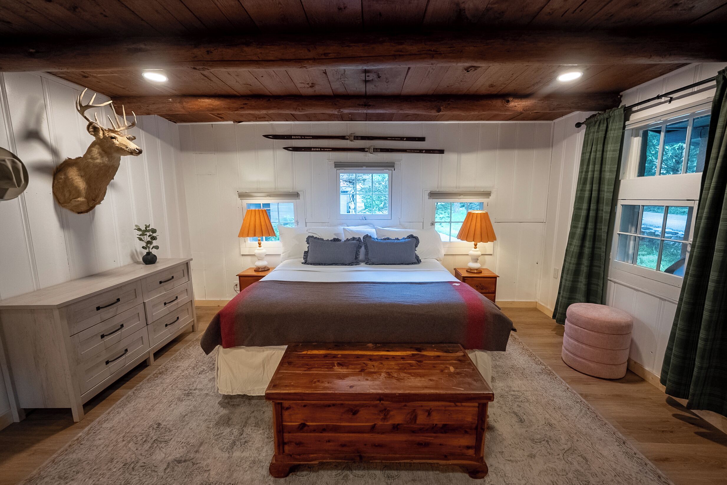 A bedroom with a large bed, wooden headboard, a mounted deer head, two bedside lamps, a window with curtains, and a wooden bench.