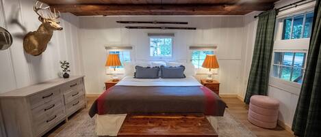 A bedroom with a large bed, wooden headboard, a mounted deer head, two bedside lamps, a window with curtains, and a wooden bench.